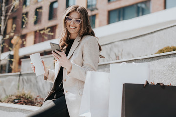 Attractive cute girl with the shopping bags and smarphone in her hand. Modern city megapolis on background. Lady making a phone call to order and shop online. Beautiful caucasian lady