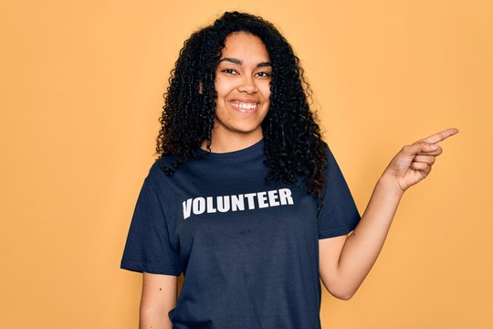 Young african american curly woman doing volunteering wearing volunteer t-shirt very happy pointing with hand and finger to the side