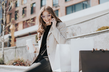 Attractive cute girl with the shopping bags and smarphone in her hand. Modern city megapolis on background. Lady making a phone call to order and shop online. Beautiful caucasian lady