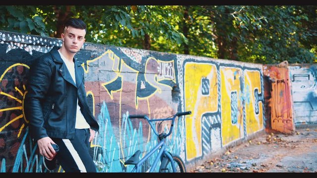 Handsome confident man is standing on graffiti background near BMX bike in a skatepark. Crop. Copy space. 4K.