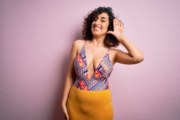 Young beautiful arab woman on vacation wearing swimsuit and sunglasses over pink background Waiving saying hello happy and smiling, friendly welcome gesture