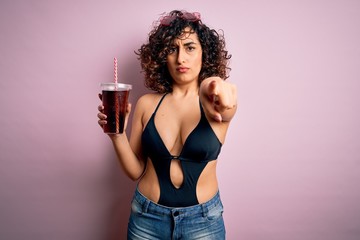 Beautiful arab woman on vacation wearing swimsuit drinking cola refreshment using straw pointing with finger to the camera and to you, hand sign, positive and confident gesture from the front