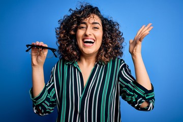 Young beautiful optical arab woman holding vision glasses over isolated blue background very happy and excited, winner expression celebrating victory screaming with big smile and raised hands