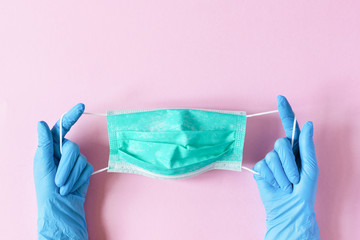 Hands in blue surgical latex gloves holding green surgical mask on pink background top view. Protection concept.