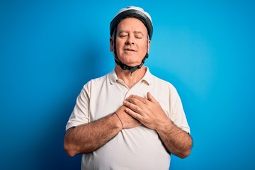 Middle age hoary cyclist man wearing bike security helmet over isolated blue background smiling with hands on chest with closed eyes and grateful gesture on face. Health concept.