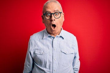Middle age handsome hoary man wearing casual striped shirt and glasses over red background afraid and shocked with surprise and amazed expression, fear and excited face.