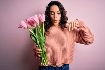 Young beautiful romantic woman with curly hair holding bouquet of pink tulips Pointing down with fingers showing advertisement, surprised face and open mouth