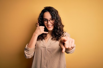 Beautiful woman with curly hair wearing striped shirt and glasses over yellow background smiling...