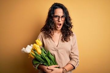 Young beautiful romantic woman with curly hair holding bouquet of yellow tulips In shock face, looking skeptical and sarcastic, surprised with open mouth