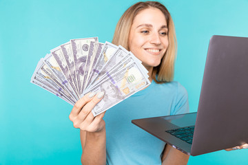 Portrait of excited satisfied girl holding money banknotes with laptop computer isolated over blue background. Portrait of a smiling young woman holding money banknotes and laptop computer in hands.