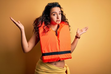 Young beautiful woman with curly hair wearing orange lifejacket over yellow background clueless and...