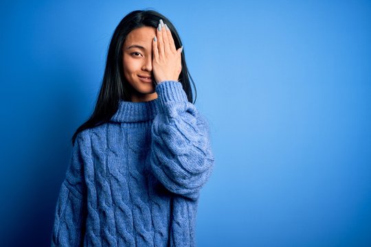 Young beautiful chinese woman wearing casual sweater over isolated blue background covering one eye with hand, confident smile on face and surprise emotion.