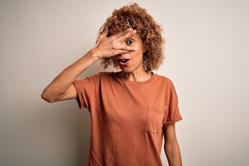 Fototapeta na wymiar Beautiful african american woman with curly hair wearing casual t-shirt over white background peeking in shock covering face and eyes with hand, looking through fingers with embarrassed expression.