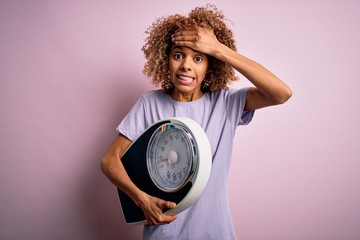 African american slim woman with curly hair holding scale over isolated pink background stressed...