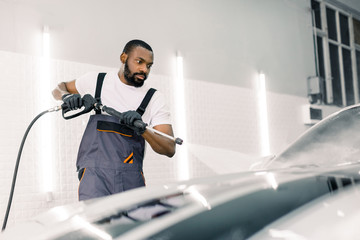 Cleaning car using high pressure water gun. Handsome young African American man worker washing...