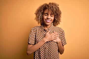 Young beautiful african american woman wearing casual shirt standing over yellow background smiling with hands on chest with closed eyes and grateful gesture on face. Health concept.