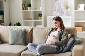 Calm attractive young pregnant woman sitting with crossed legs on sofa and embracing her belly