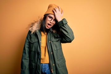 Young brunette woman wearing glasses and winter coat with hat over yellow isolated background...