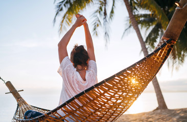 Young woman awaking and meeting morning sunrise sunlight sitting in a hammock and lazy stretching her body raising arms up on the sandy beach under the palm trees. Calm exotic places vacation concept