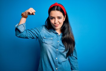 Young brunette woman wearing casual denim shirt over blue isolated background Strong person showing arm muscle, confident and proud of power