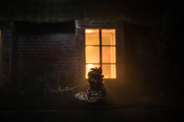 Fototapeta na wymiar Horror concept. Little baby standing in the yard near a window of old creepy house. Scary baby silhouette in dark. A realistic dollhouse at night. Creative artwork decoration.