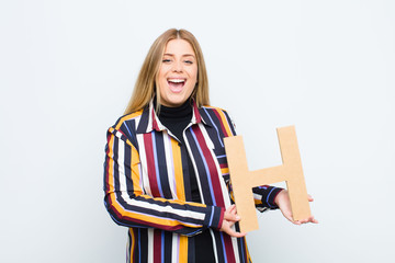 young pretty blonde woman excited, happy, joyful, holding the letter H of the alphabet to form a...
