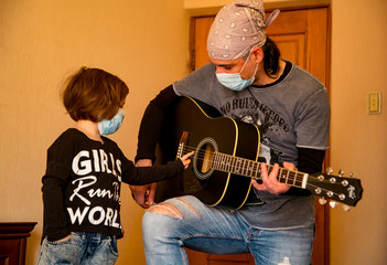a little girl with her dad in medical masks, and rock-style, learning to play guitar, at home during quarantine, due to the covid-19 pandemic