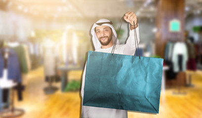 Handsome young Arab man in Kandura showing his shopping bags and smiling while standing in mall..