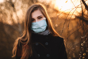 Portrait of happy cute young woman wearing protective face medical mask standing in park at sunset during spring