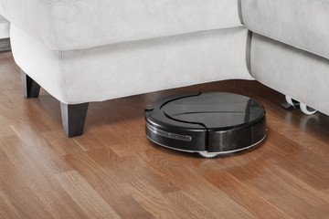 black Robotic vacuum cleaner runs under sofa. Robot controlled by voice commands to direct cleaning. Modern smart cleaning technology housekeeping