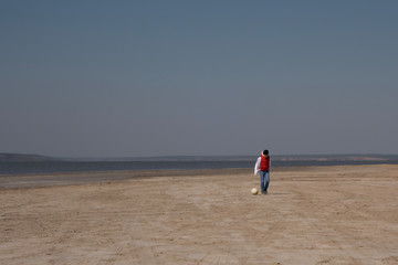 A boy of 10 years old in a white sweatshirt and orange vest plays football on a deserted beach in solitude.