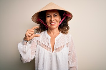 Middle age brunette woman wearing asian traditional conical hat over white background smiling and confident gesturing with hand doing small size sign with fingers looking and the camera. Measure