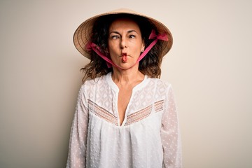 Middle age brunette woman wearing asian traditional conical hat over white background making fish face with lips, crazy and comical gesture. Funny expression.