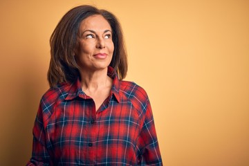 Middle age beautiful woman wearing casual shirt standing over isolated yellow background smiling looking to the side and staring away thinking.