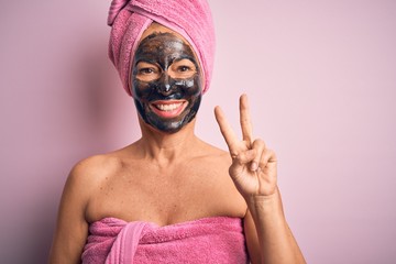 Middle age brunette woman wearing beauty black face mask over isolated pink background smiling looking to the camera showing fingers doing victory sign. Number two.