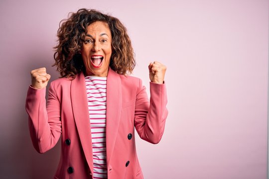 Middle age beautiful businesswoman wearing elegant jacket over isolated pink background celebrating surprised and amazed for success with arms raised and open eyes. Winner concept.