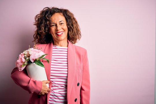Middle age beautiful woman holding vase with flowers over isolated pink background with a happy face standing and smiling with a confident smile showing teeth