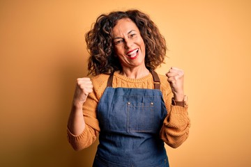 Middle age beautiful baker woman wearing apron standing over isolated yellow background celebrating surprised and amazed for success with arms raised and open eyes. Winner concept.
