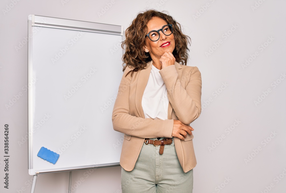 Wall mural Middle age senior business woman standing on seminar presentation by magnectic blackboard with hand on chin thinking about question, pensive expression. Smiling and thoughtful face. Doubt concept.