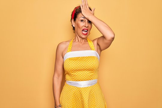 Middle age senior pin up woman wearing 50s style retro dress over yellow background surprised with hand on head for mistake, remember error. Forgot, bad memory concept.