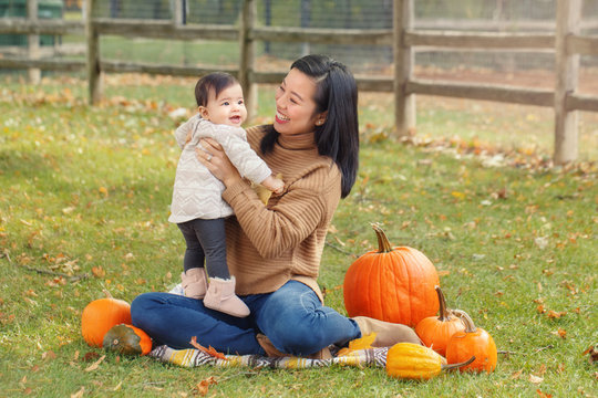 Happy Smiling Asian Chinese Mother With Cute Adorable Baby. Mom And A Daughter Girl Family Sitting In Autumn Fall Park Outdoor With Pumpkins. Halloween Or Thanksgiving Seasonal Concept.