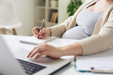 Close-up of unrecognizable pregnant woman sitting at table and using laptop while writing down...