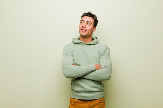 Young Arabian Man Feeling Happy, Proud And Hopeful, Wondering Or Thinking, Looking Up To Copy Space With Crossed Arms Against Flat Wall