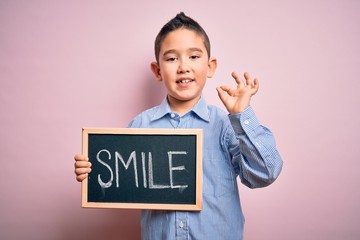 Young little boy kid showing blackboard with smile word as happy message over pink background doing ok sign with fingers, excellent symbol