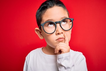 Young little smart boy kid wearing nerd glasses over red isolated background serious face thinking about question, very confused idea