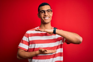 Young handsome african american man wearing casual striped t-shirt and glasses gesturing with hands showing big and large size sign, measure symbol. Smiling looking at the camera. Measuring concept.