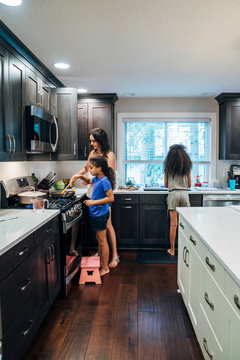 Mom and daughter cooking meal together in home kitchen, while girl washes dishes in back ground