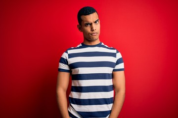 Handsome african american man wearing casual striped t-shirt standing over red background skeptic...
