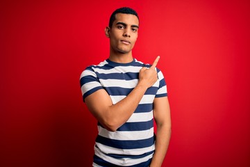 Fototapeta premium Handsome african american man wearing casual striped t-shirt standing over red background Pointing with hand finger to the side showing advertisement, serious and calm face