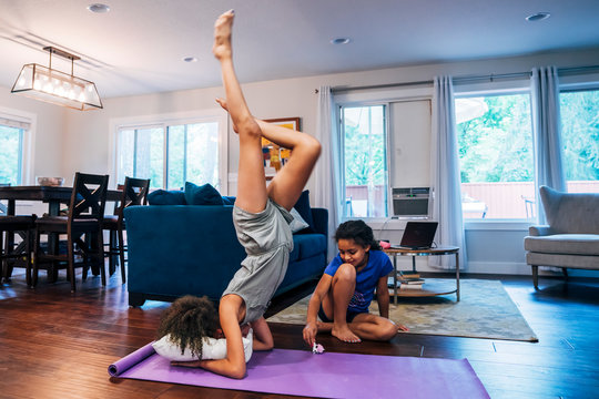 Girl doing headstand on pillow and yoga mat while sister plays in background